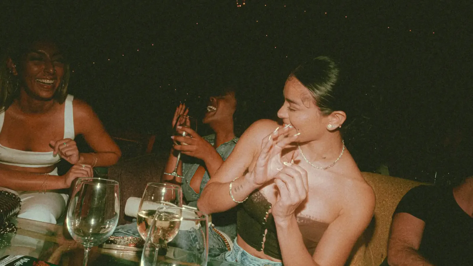 Trois amies éclatent de rire autour d’une table, verres de vin à la main, lors d’une soirée entre inconnus